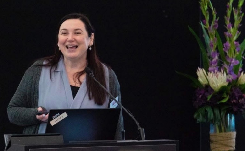 A woman with long dark hair, smiling warmly, stands behind a dark wooden lectern during a presentation. She wears a light blue blouse under a grey cardigan. A laptop and a microphone are positioned on the lectern in front of her. To her right is a large, vibrant floral arrangement featuring purple gladioli and white proteas in a glass vase. The background consists of a plain black partition.