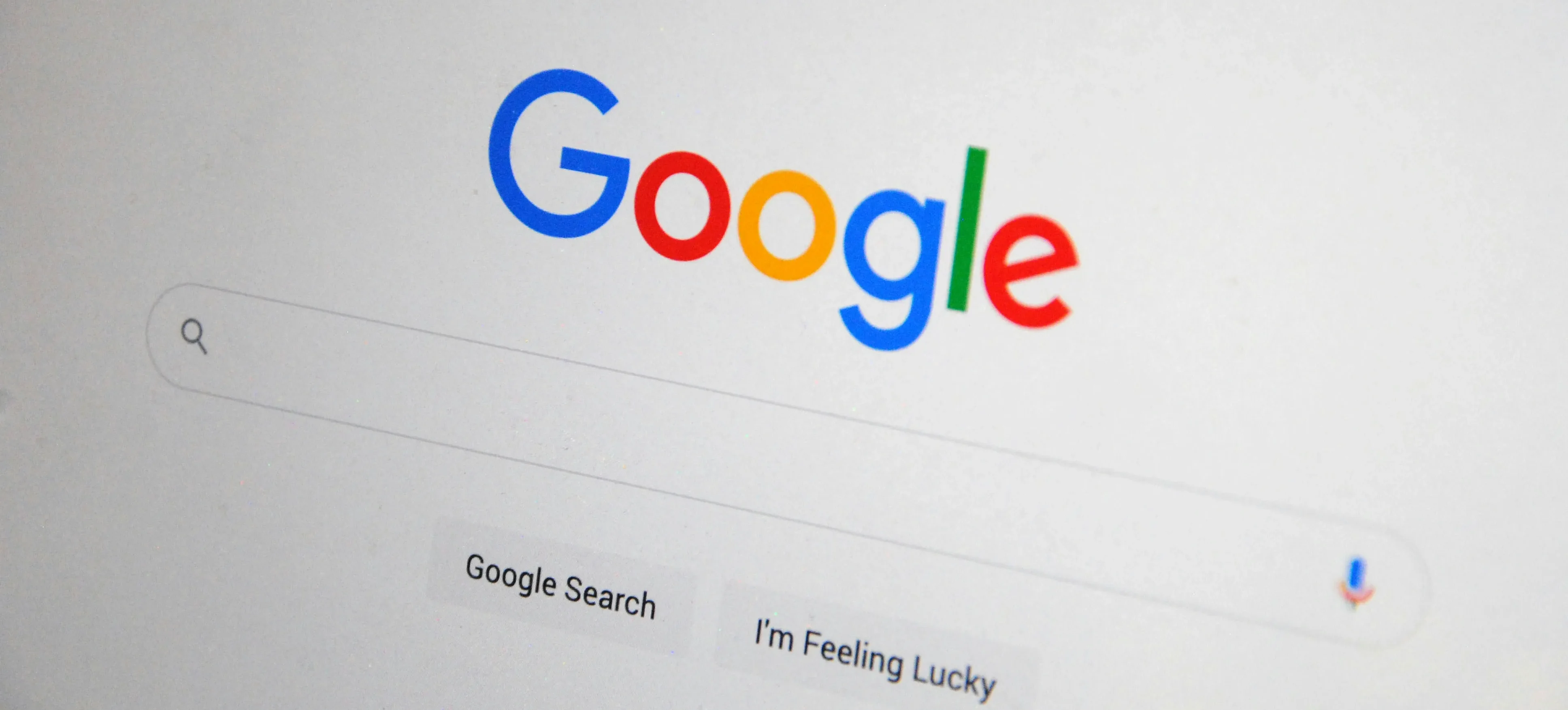 A close-up photograph of a laptop or monitor screen displaying the Google homepage, showing the colourful Google logo, an empty search bar, and the "Google Search" and "I'm Feeling Lucky" buttons on a white background.